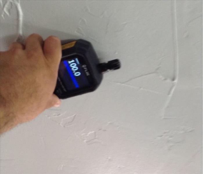 Hand holding moisture meter that reads 100, high moisture, against drywall that looks to be clean with no signs of moisture